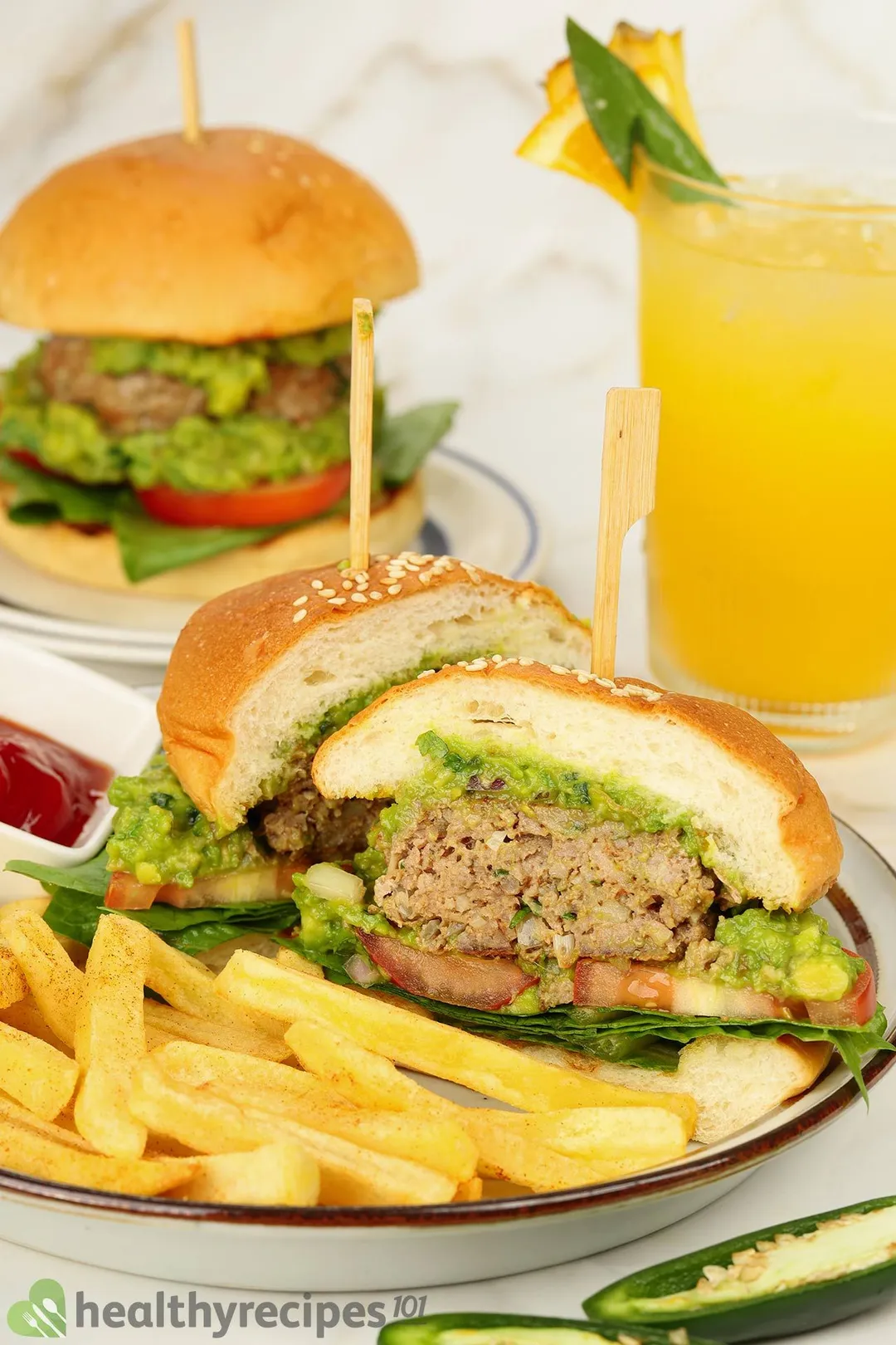 front shot of a half burger on a plate with french fries and yellow glass juice, a burger in background