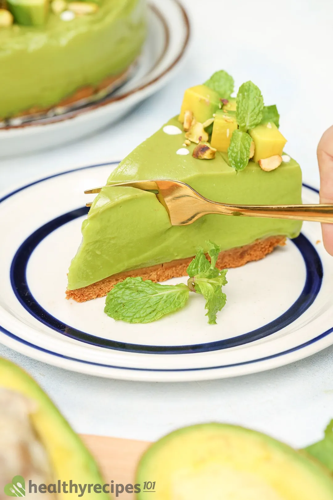 A golden fork cutting into a slice of avocado cheesecake garnished with green mints and avocado cubes