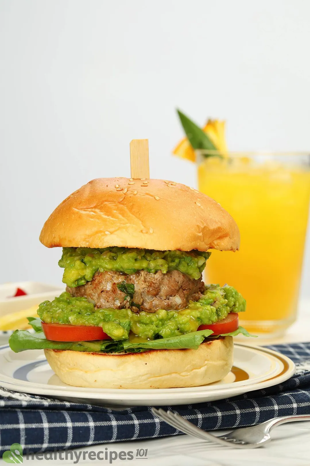 a plate with an avocado burger on it, a yellow glass juice in background