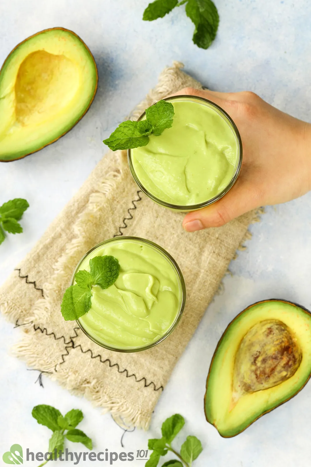 two glasses of avocado smoothie on table, a hand holding one glass