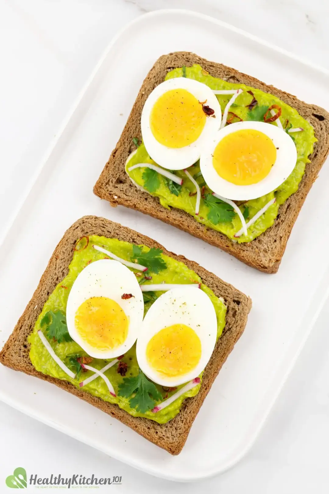 Two slices of brown toast topped with avocado smear, boiled egg slices, cilantro sprigs, and other garnishes