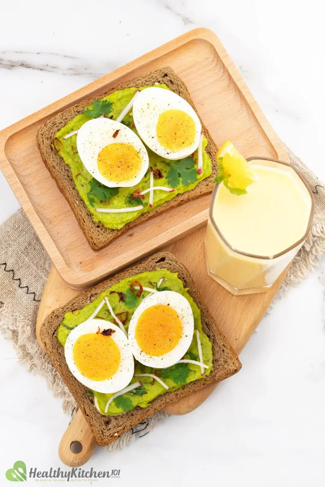 Two avocado toast slices put next to each other on wooden boards, a cloth, and a glass of mango smoothie