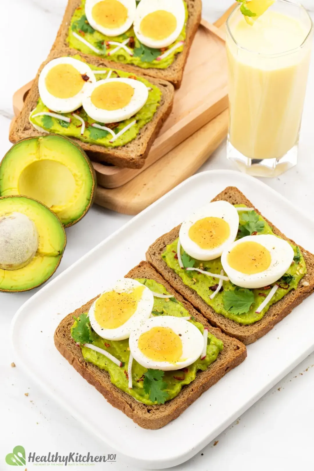 Four slices of avocado toast and boiled eggs plated together with halved avocado and a glass of mango smoothie