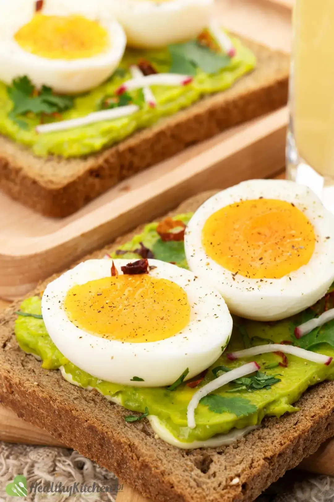 A slice of avocado toast topped with 2 slices of boiled eggs and garnishes, with another toast in the background