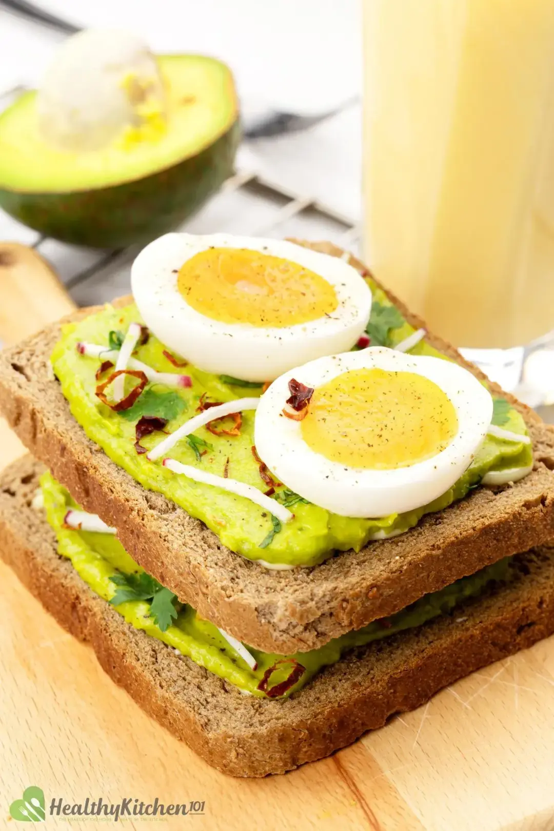 Two slices of avocado toast with boiled eggs put on top of one another, with a smoothie and an avocado in the background