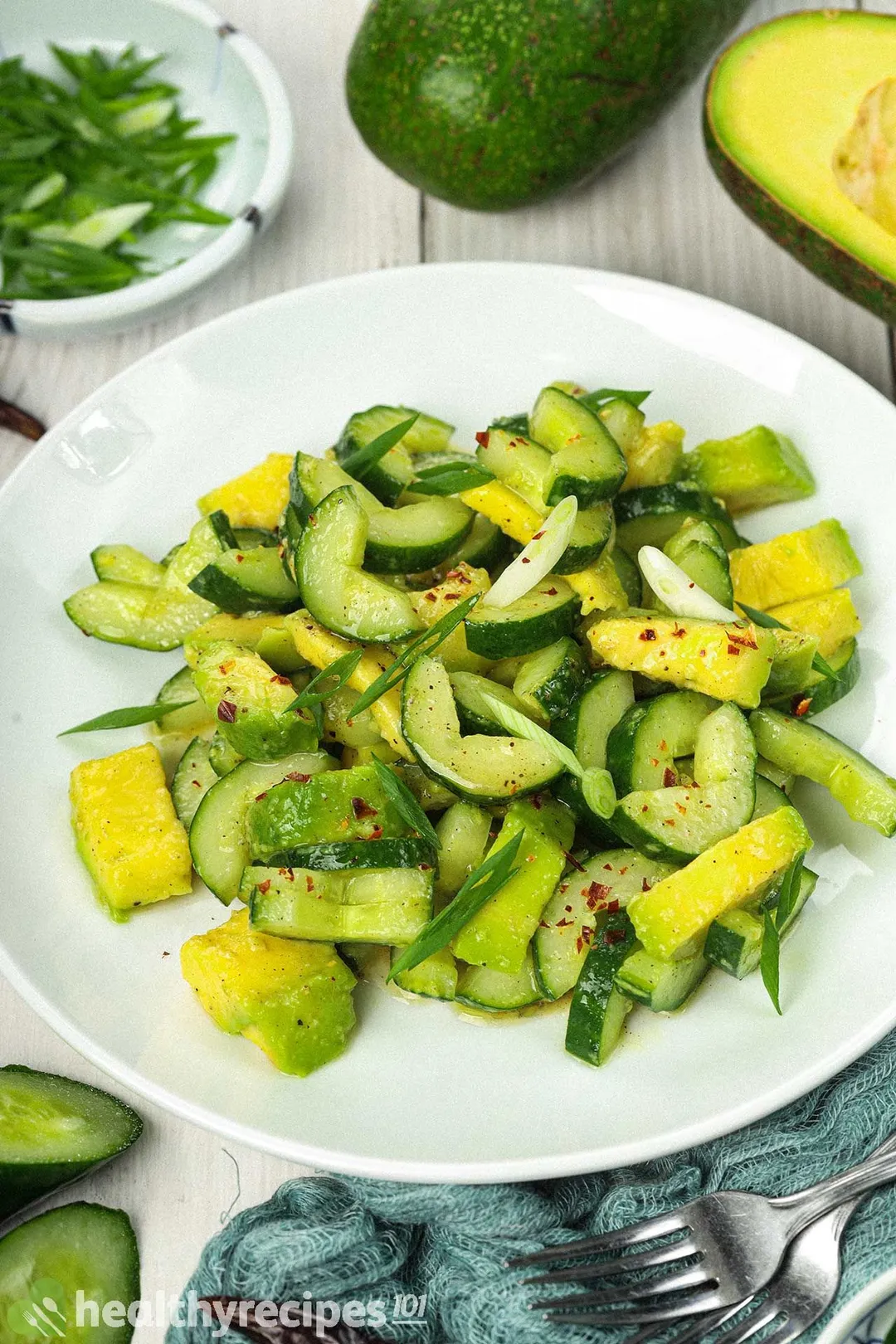 A round white plate containing cucumber avocado salad laid near two forks and a few cucumber slices
