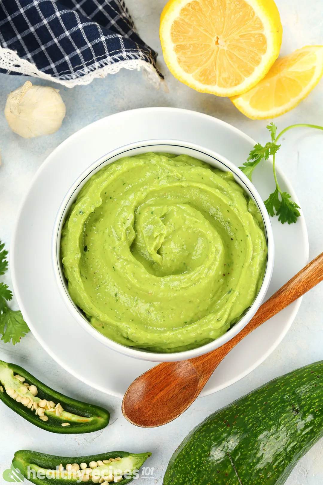 a bowl of avocado sauce on a plate with a wooden spoon, decorated with avocado, half jalapeno, lemon and garlic