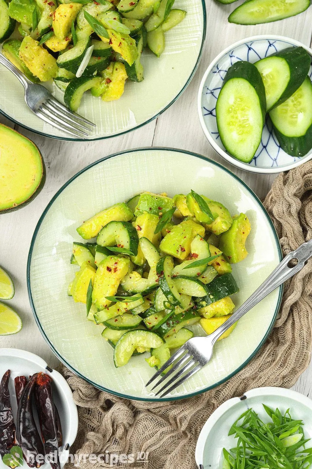 Plates of cucumber avocado salad laid near a fork, chopped scallions, dried chili peppers, and cucumber slices