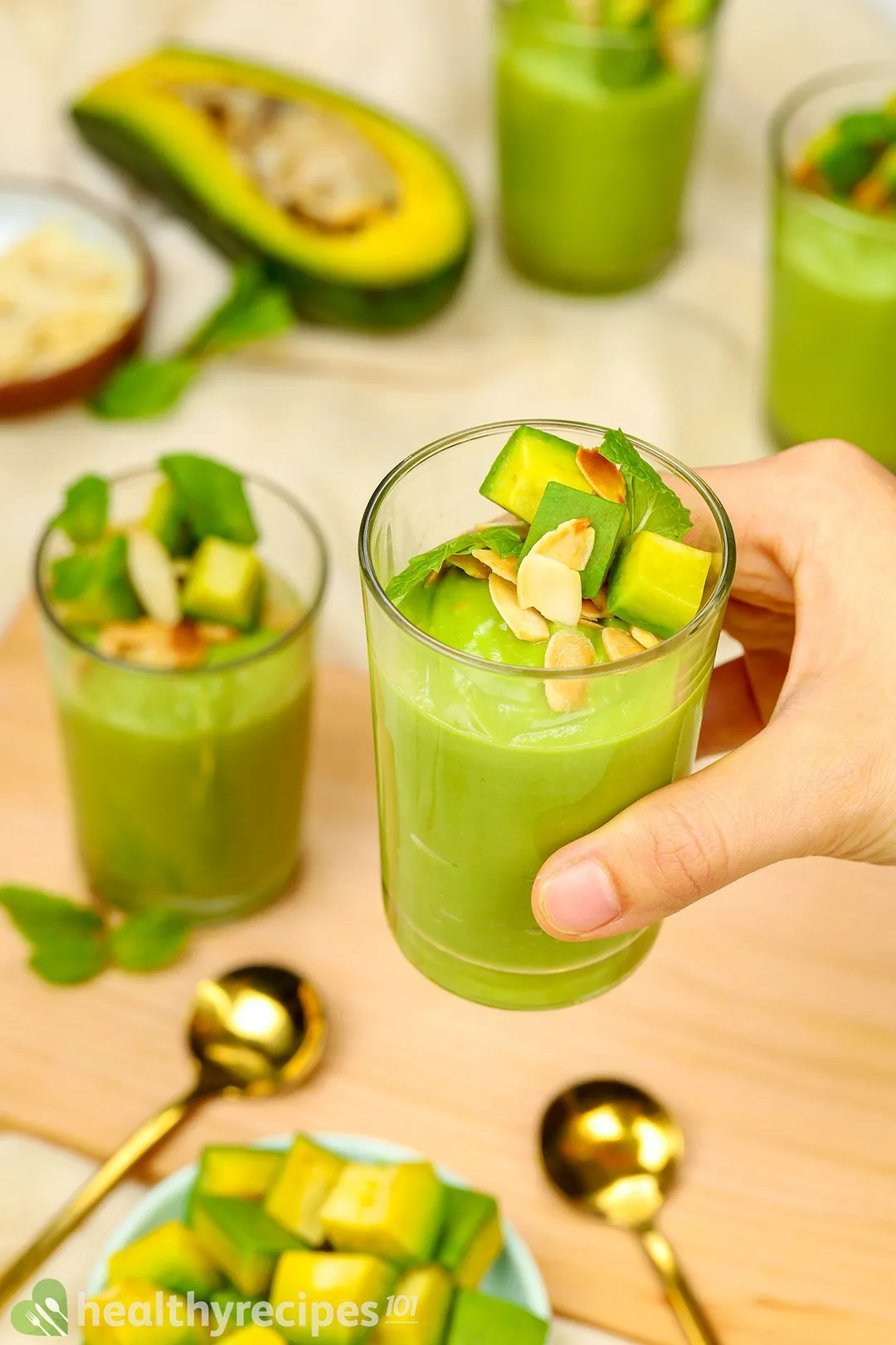 a hand holding an avocado mouse glass with three glasses and half avocado, two golden spoon on background
