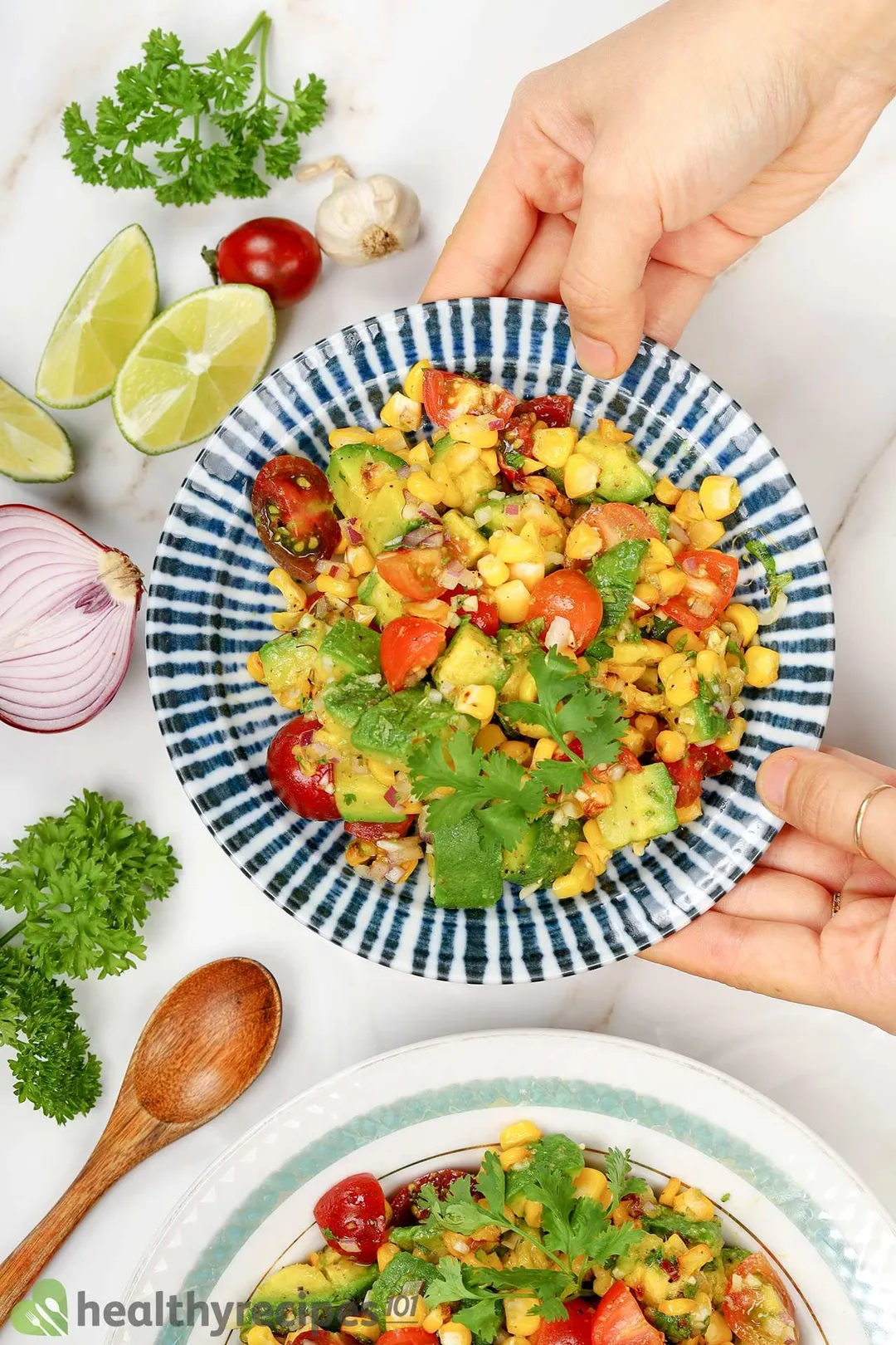 two hands holding a plate of avocado cubes, corn, tomato cubes and coriander, decorated by half onion, a wooden spoon, slices lime tomato and garlic