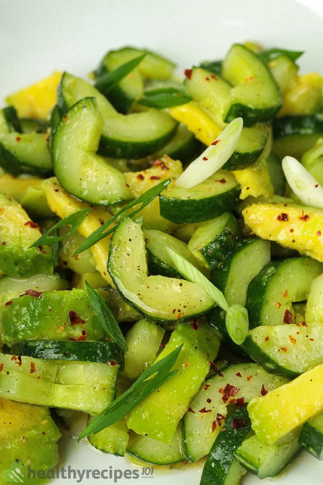 A close-up shot of half-moon sliced cucumbers, avocado cubes, dried pepper flakes, and chopped scallions