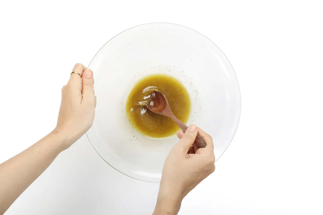 A hand steadying a large glass bowl while the other hand is stirring a dark brown sauce in it with a wooden spoon