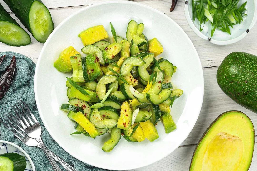 A round white plate containing cucumber avocado salad laid near sliced cucumbers, avocado halves, and two forks