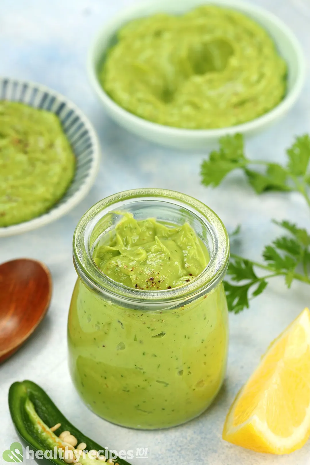 front shot of a jar of avocado sauce, surrounded by lemon wedge, half jalapeno, two bowl of green sauce