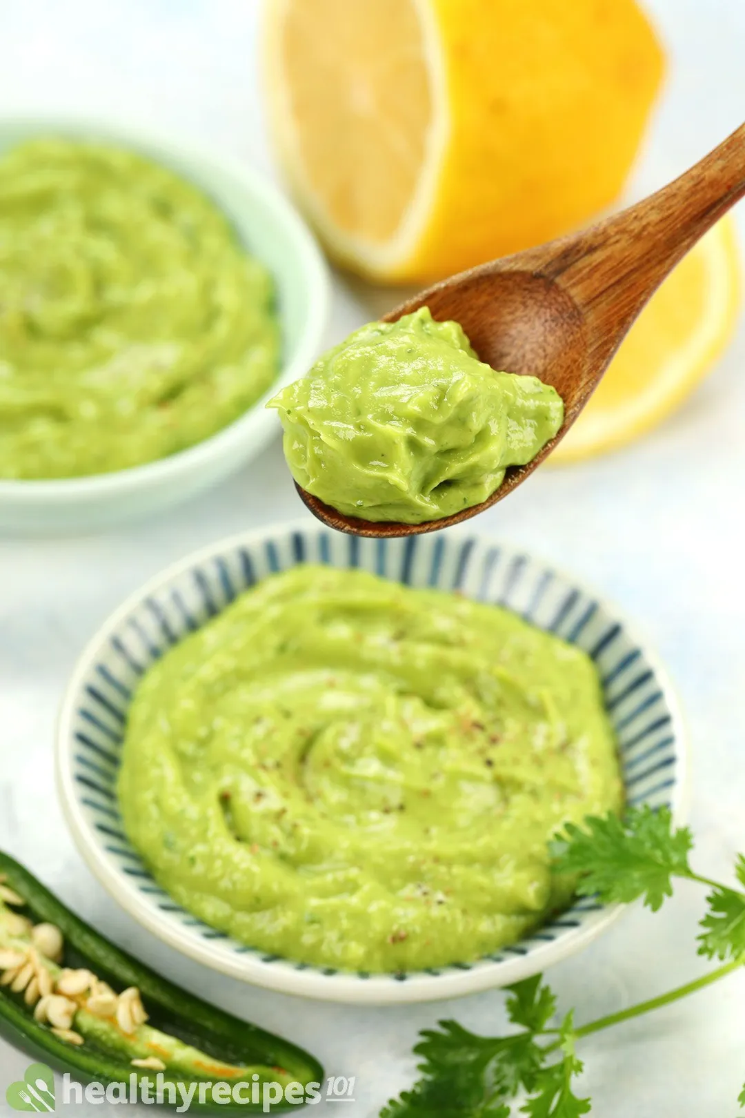 a wooden spoon of avocado sauce on top of a bowl of sauce, half lemon and a bowl of sauce in background 