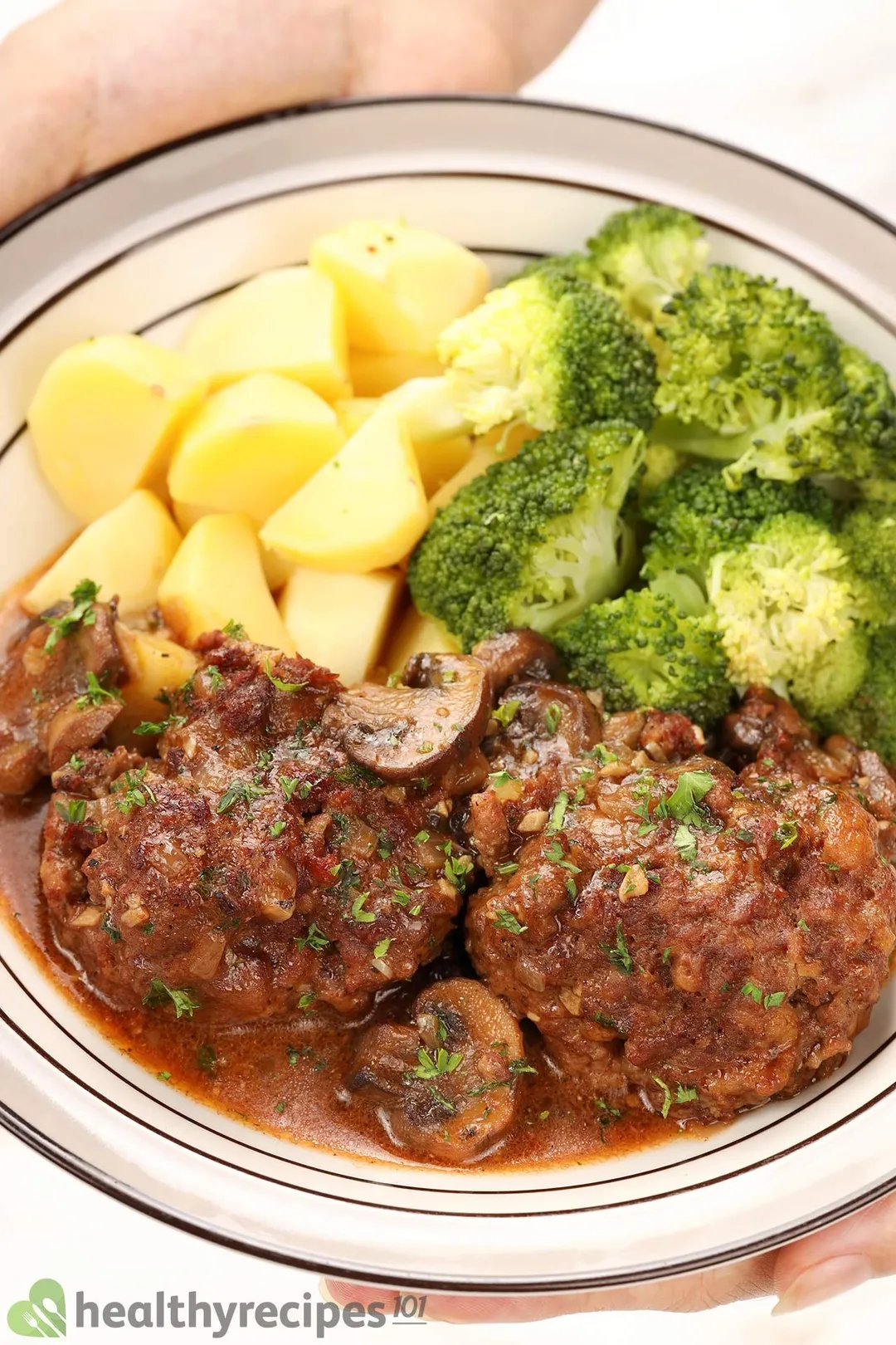 hand holding a plate of cooked beef with mushroom, cubed potatoes and broccoli