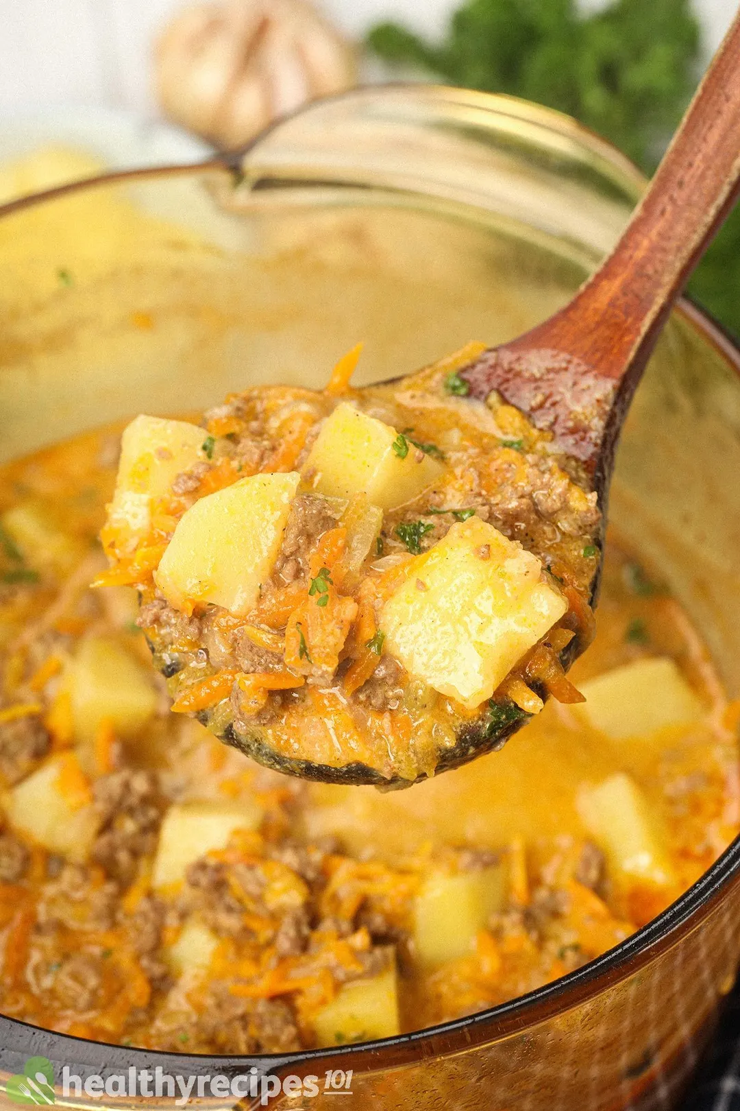 A wooden ladle filled with cooked potato cubes, julienned carrots, and ground beef held over a glass saucepan filled with cheeseburger soup