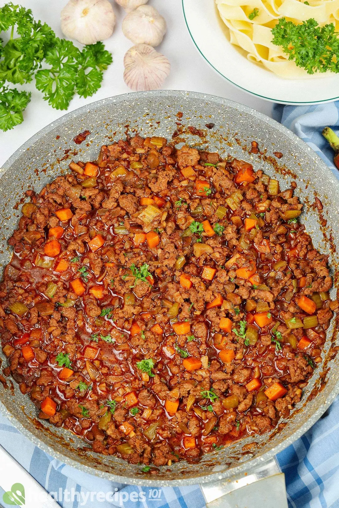 a nonstick skillet of cooked ground beef and veggies, decorated with garlic and a plate of pasta, parsley