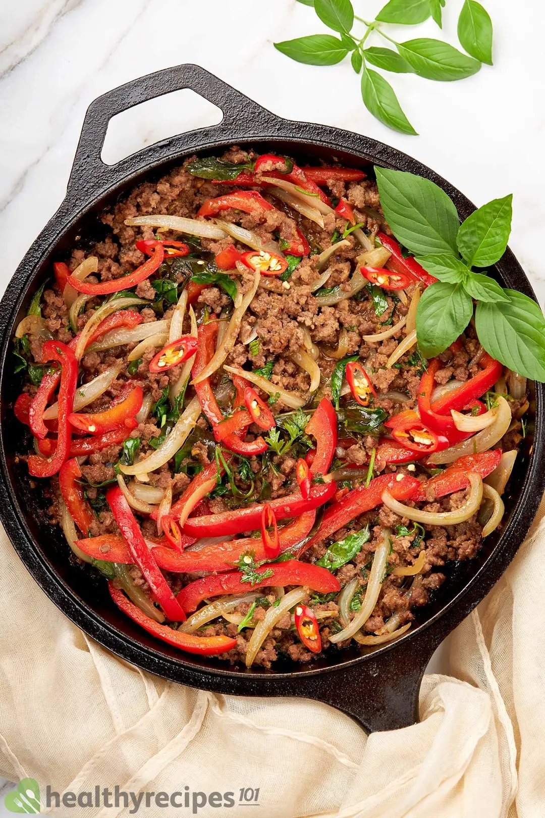 a cast iron skillet of cooked ground beef with sliced onion, red bell pepper and chopped chili decorated by basil