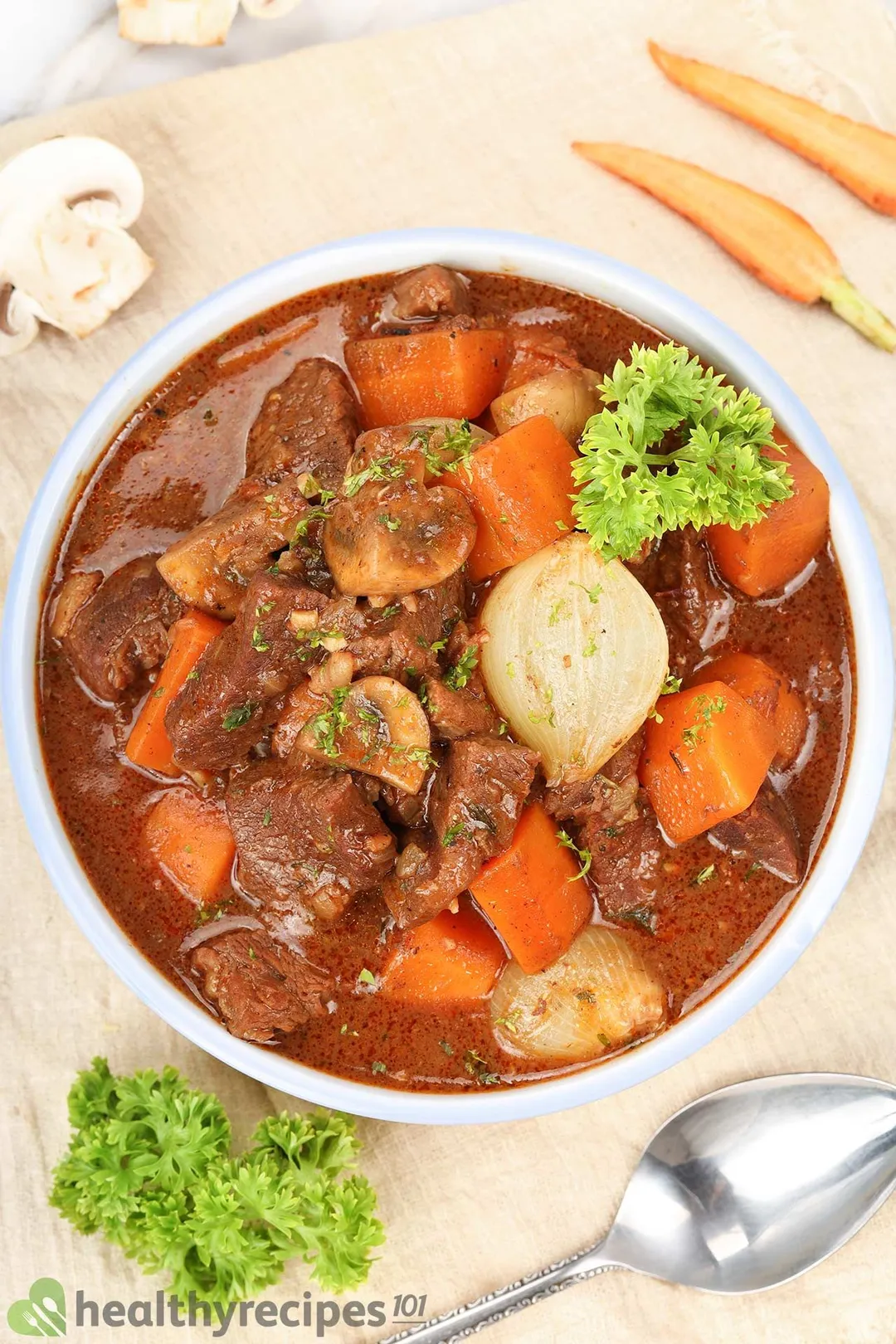 top view of a bowl of cooked cubed beef chuck with cubed carrot, onion, garnished by half carrot, half mushroom, parsley and a spoon