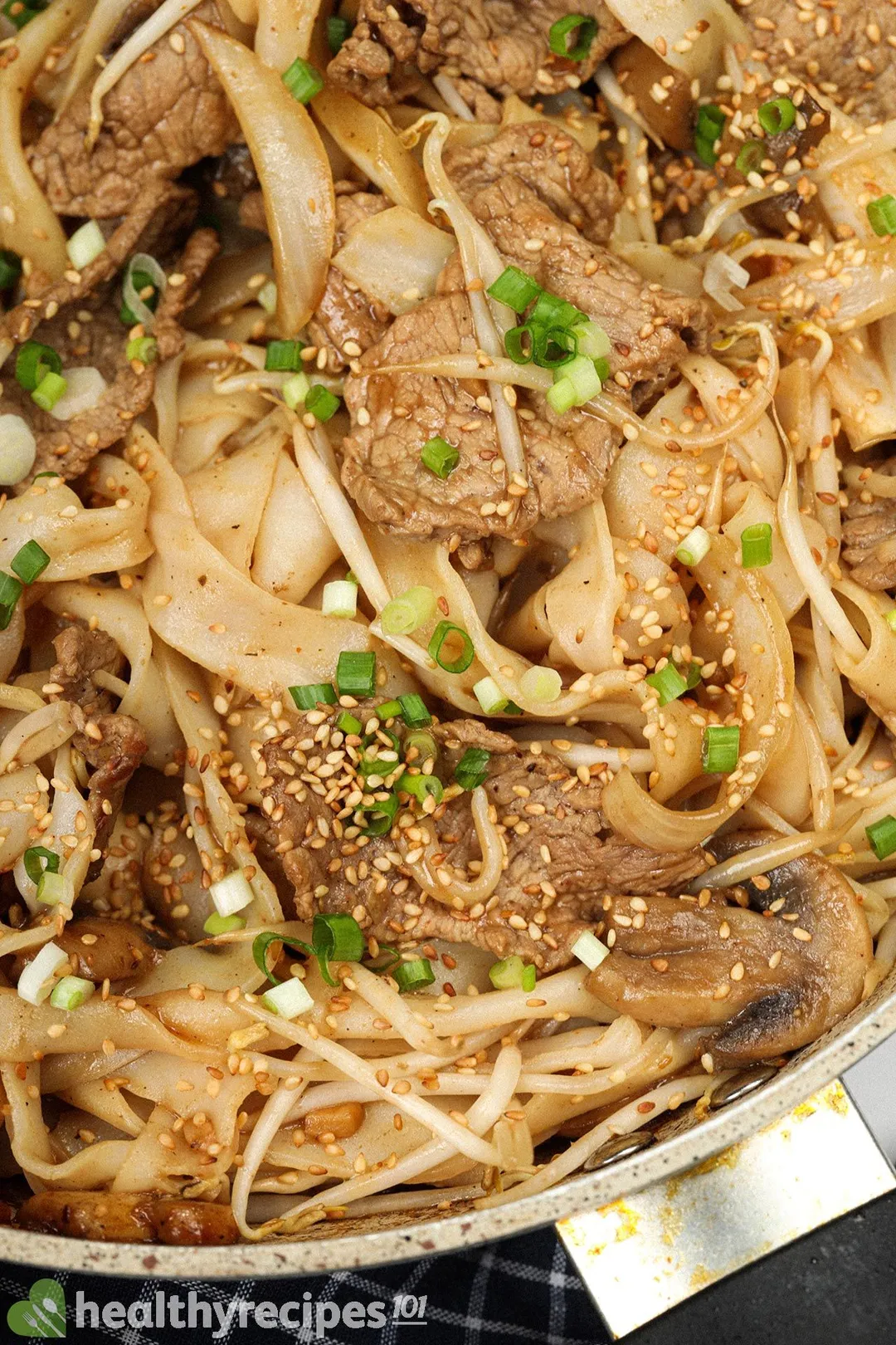 close-up shot of skillet of cooked noodle and sliced beef with sesame seed, chopped scallion, mung bean sprouts