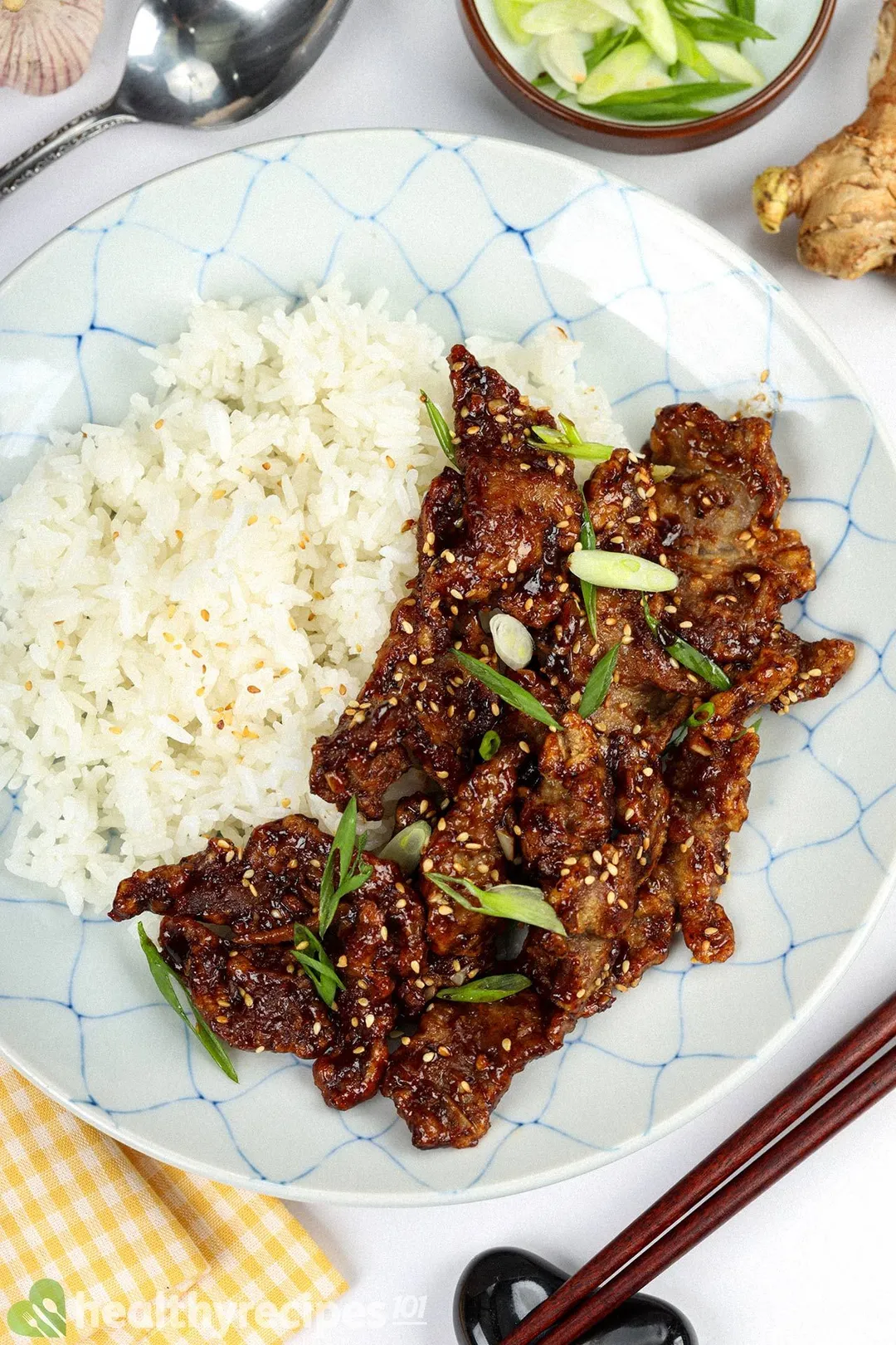 a plate of cooked ginger beef with rice, surrounded by a spoon and pair of chopstick, ginger, garlic, chopped scallion