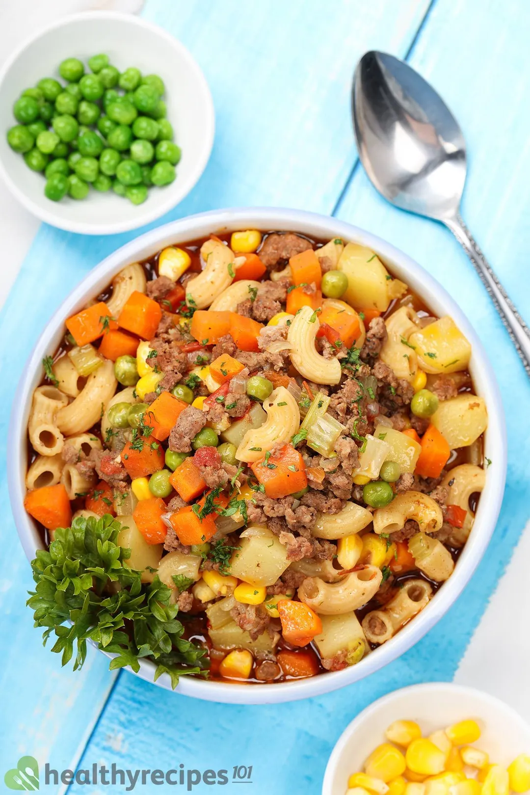top view of a bowl of cooked ground beef with pasta, carrot and potatoes cubed, green bean, corn garnished with a spoon, a small bowl of green bean, corn