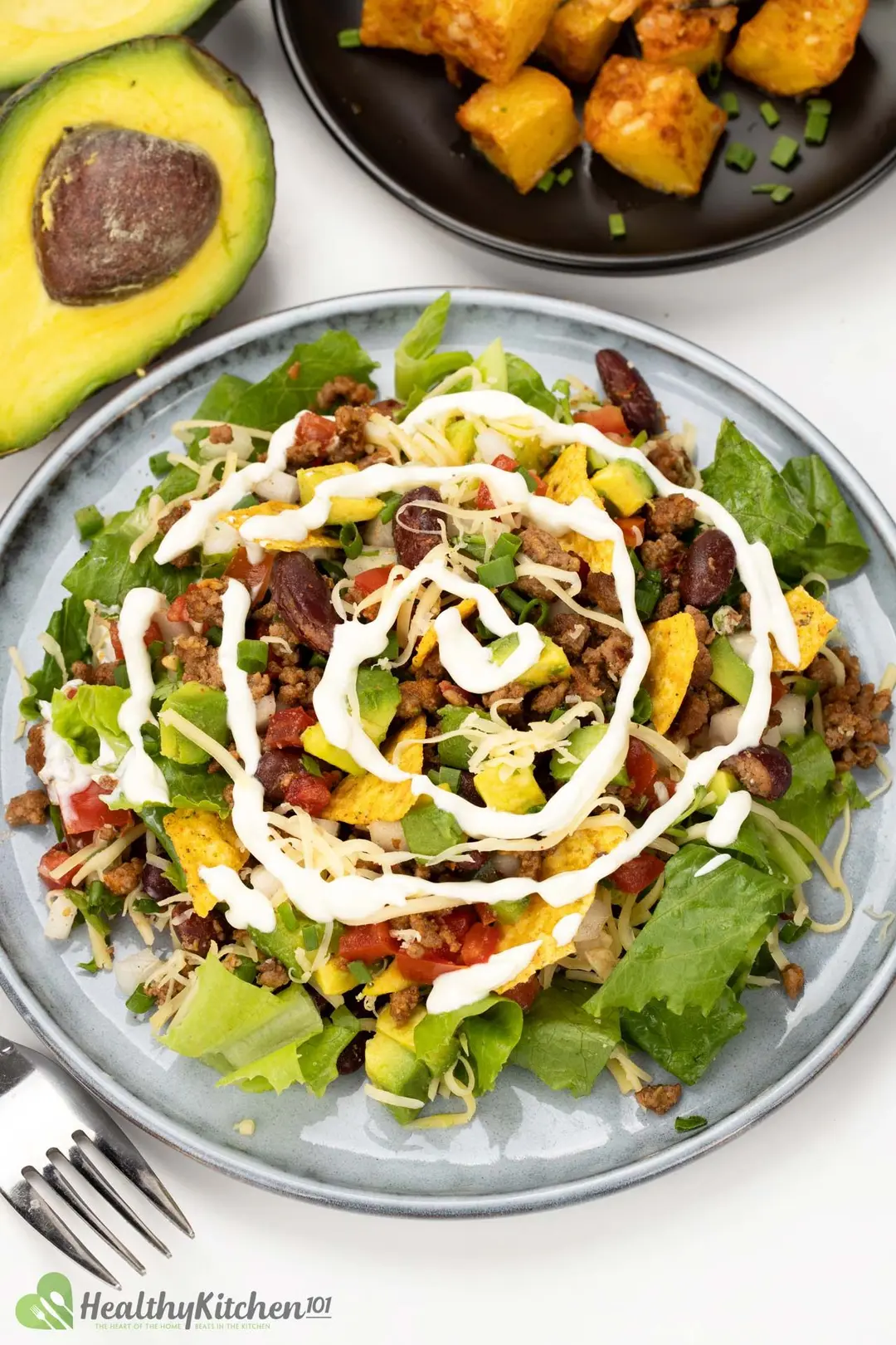 a plate of taco salad with white dressing decorated by half avocado, a plate of cooked potato cubed and a fork