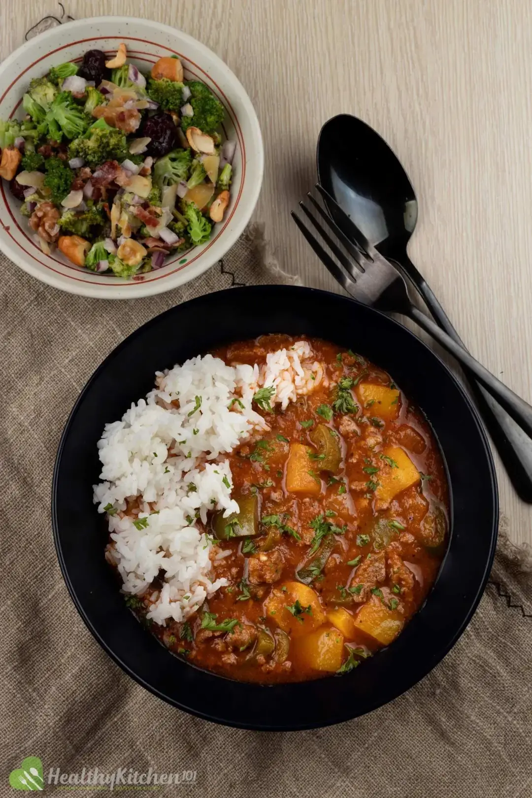 A bowl of stuffed pepper soup and cooked white rice placed on the side, next to utensils and a salad
