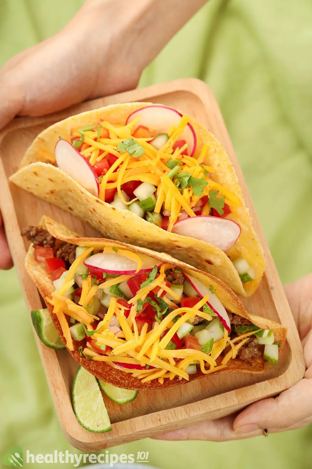 two hand holding a wooden tray of two tortillas with cooked beef and veggies in a green background