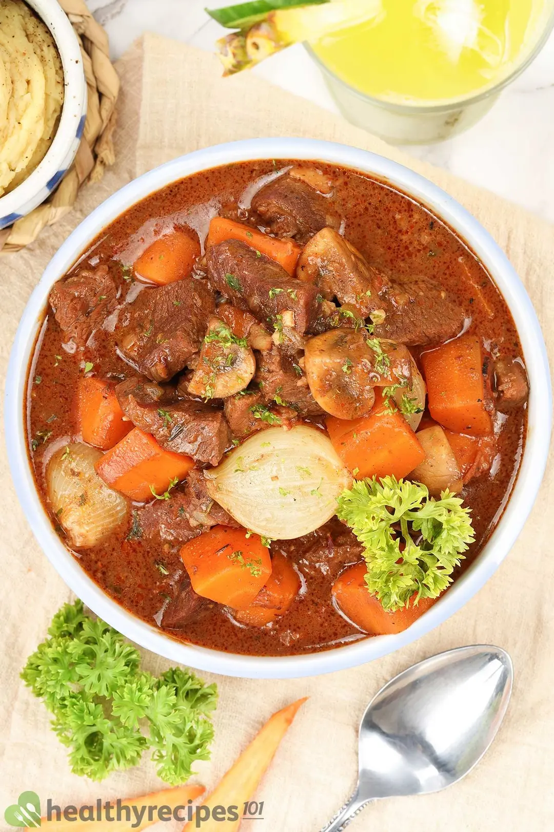 A high-angle shot of a beef stew bowl consisting of a dark brown liquid, large beef cubes, carrot cubes, potato cubes, and fresh parsley laid near various decorative items
