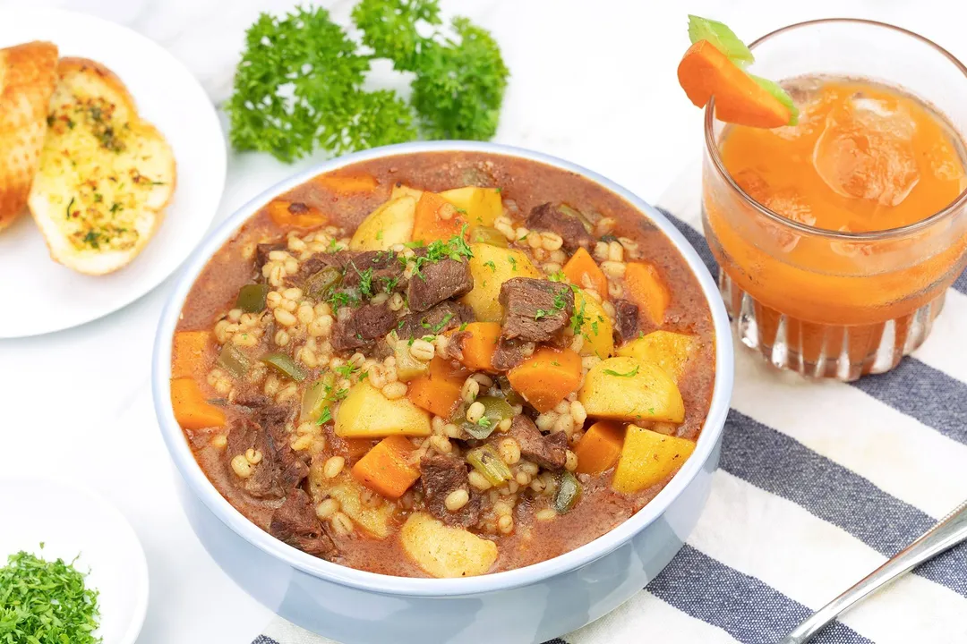 a bowl of beef barley soup next to a glass of carrot juice and a plate of garlic bread, decorated with parsley