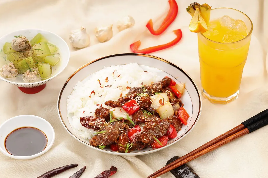 a plate of beijing beef and rice next to a glass of pineapple juice and a bowl of winter melon soup and a small bow of sauce