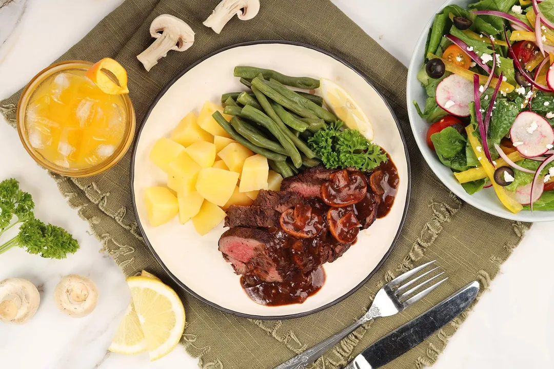 a plate of beef tenderloin with potato cubed and green beans, decorated with a glass of orange juice and a plate of salad
