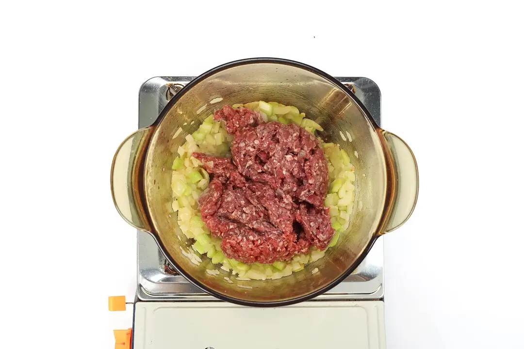 A glass saucepan containing diced vegetables and ground beef cooking on a portable gas stove