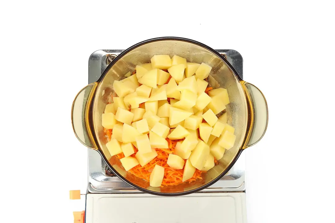 A glass saucepan containing potato cubes and julienned carrots cooking on a portable gas stove