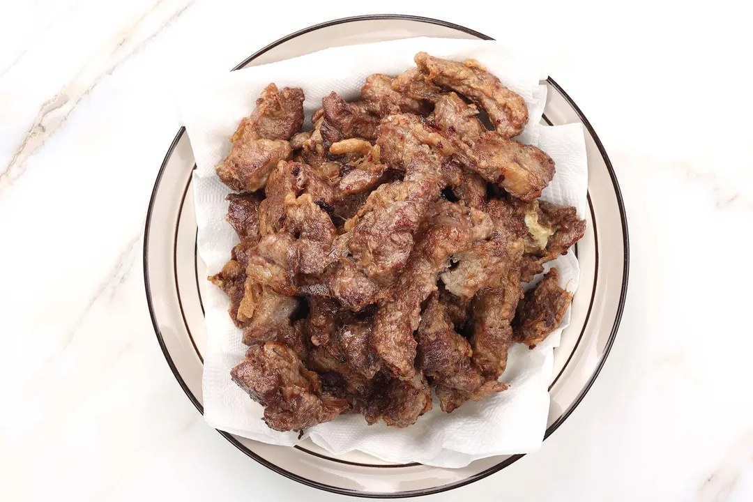 cooked beef on a plate lined with paper towels to catch excess oil from the beef.