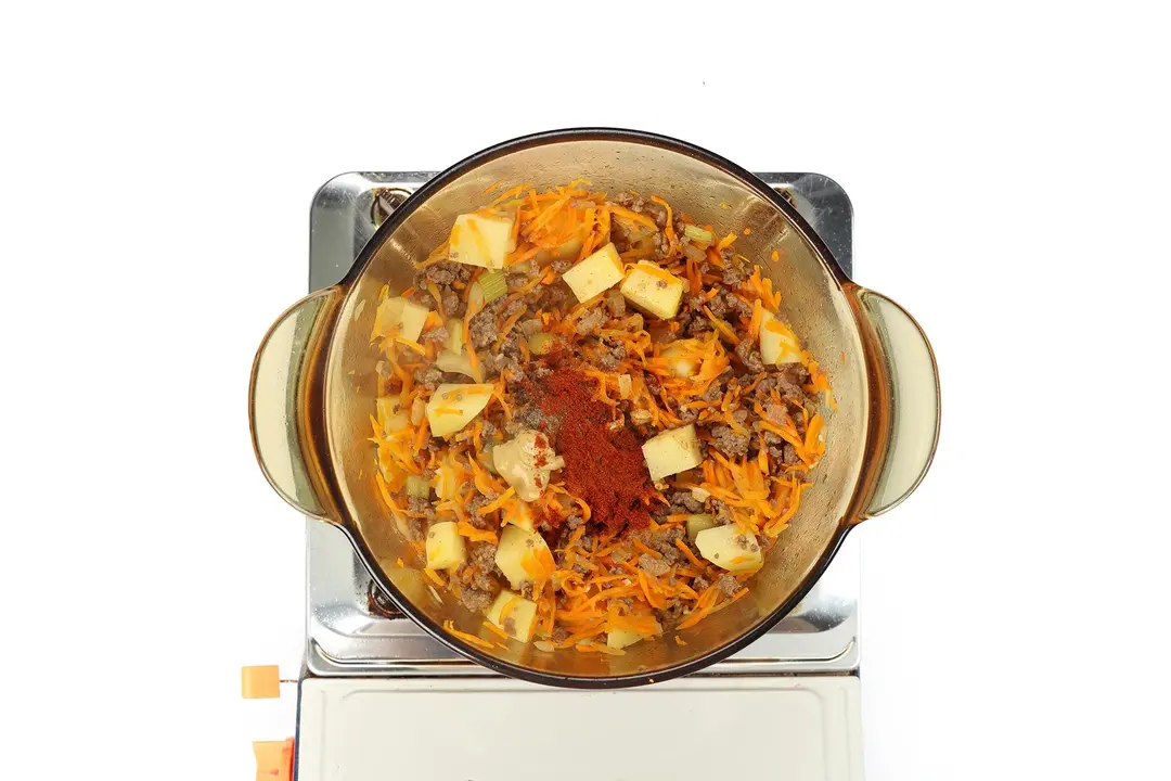 A glass saucepan containing potato cubes, ground beef, julienned carrots, and paprika cooking on a portable gas stove