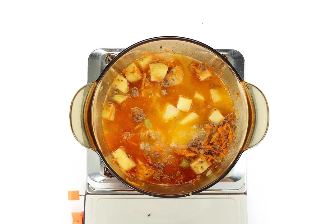 A glass saucepan cooking a soup featuring potato cubes, ground beef, and julienned carrots on a portable gas stove