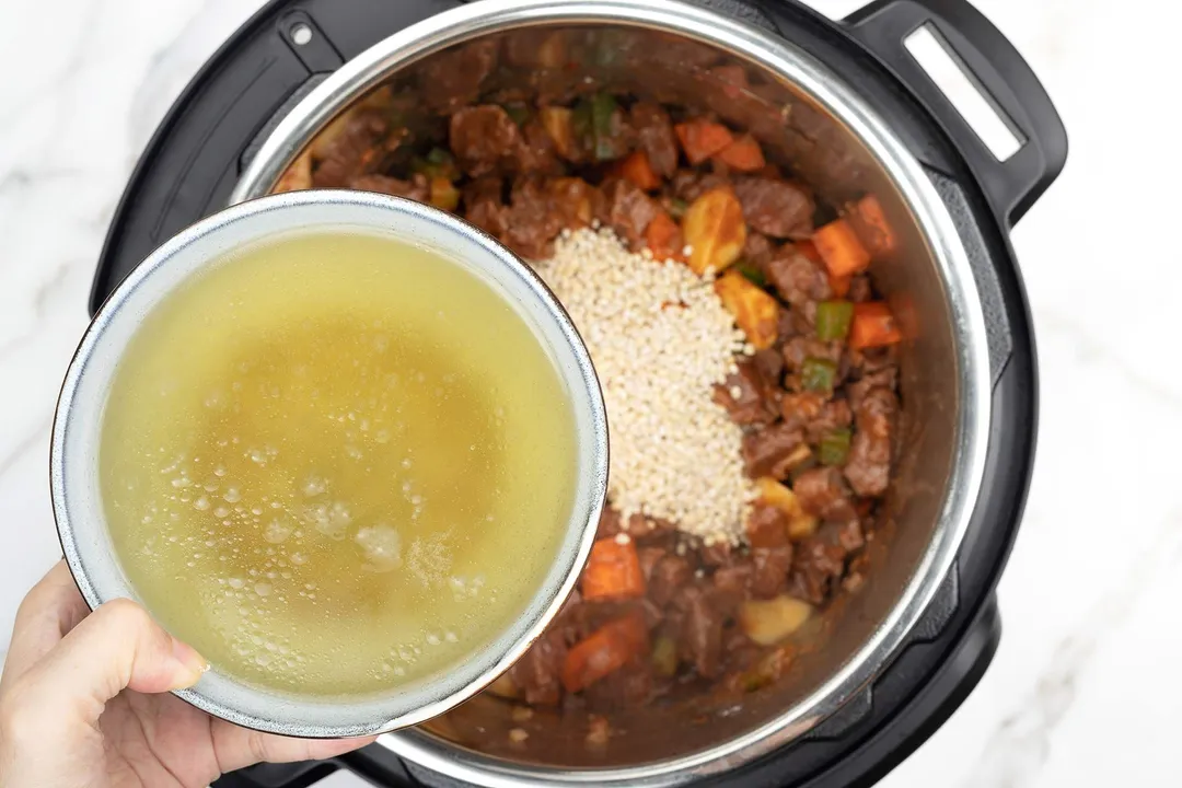 a hand holding a bowl of chicken broth on top of an instant pot with beef and barley cooking in it