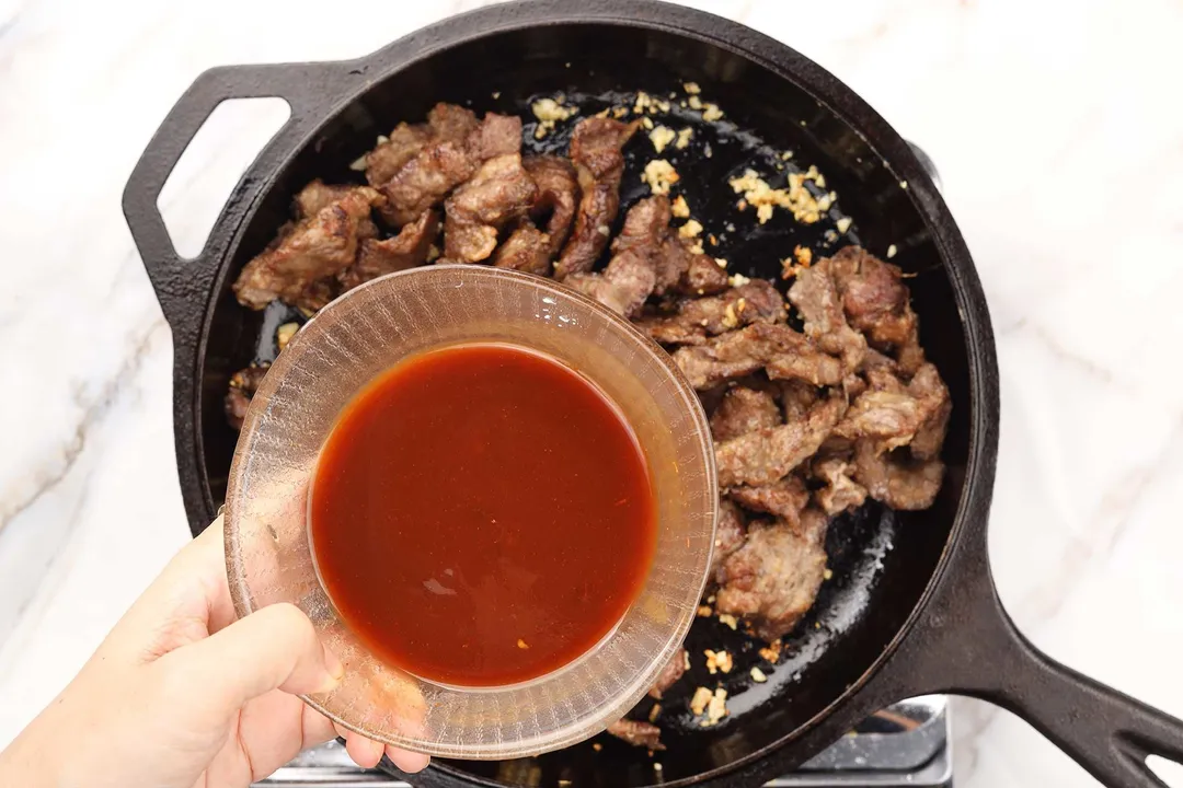 a hand holding a glass bowl of sauce on top of a cast iron skillet of cooked beef chunk