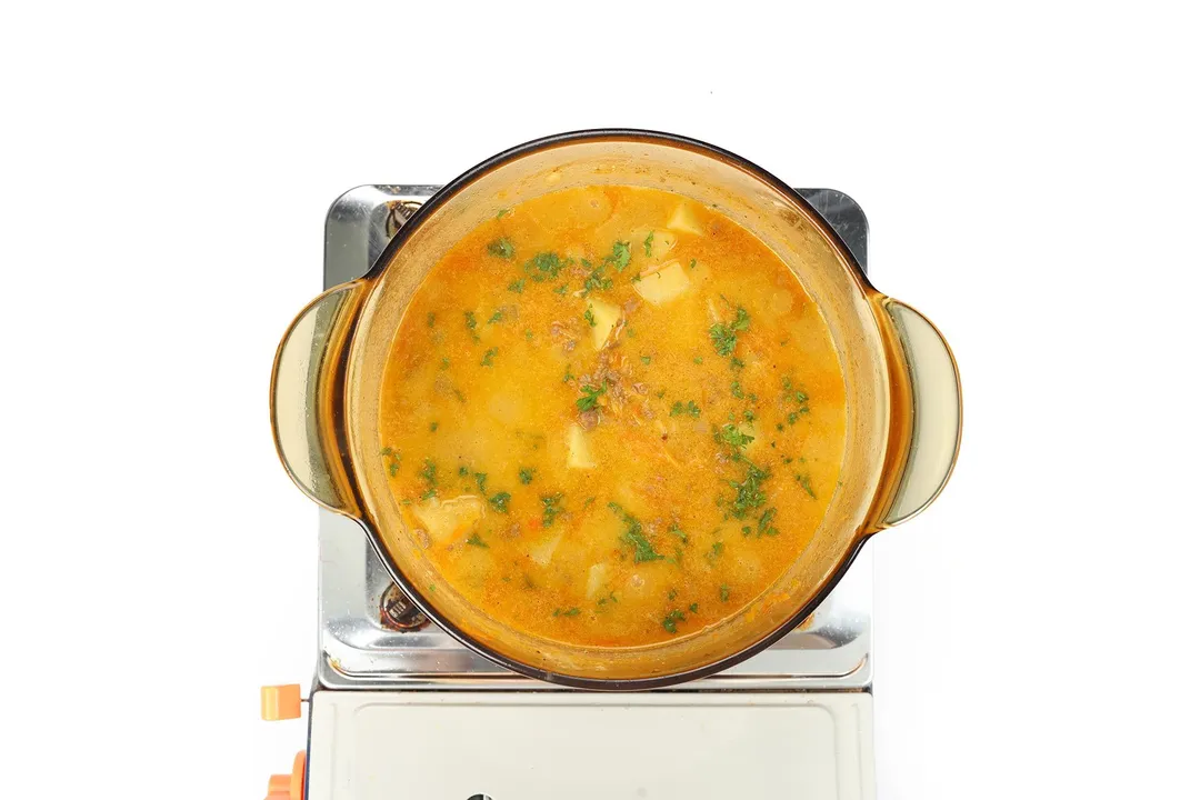 A glass saucepan cooking a bright yellow soup with chopped parsley on the surface on a portable gas stove