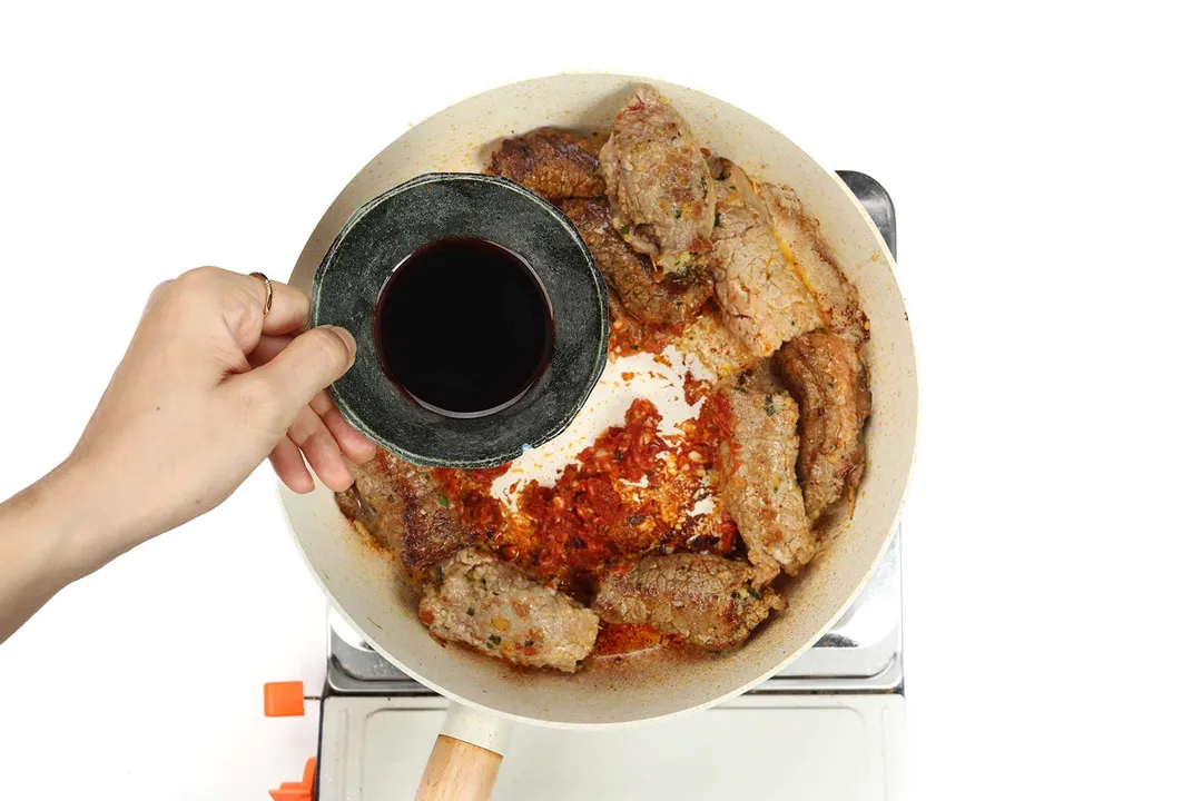 hand holds a bowl of red wine on top of a skillet of cooked beef