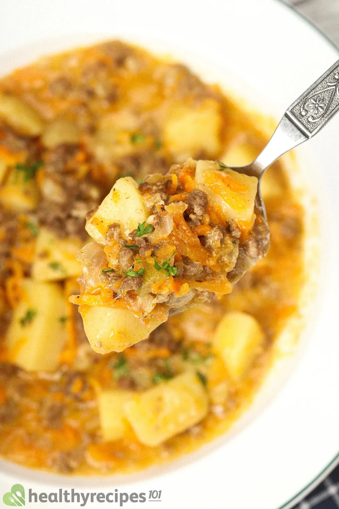 A spoon scooping up cooked potato cubes, ground beef, and julienned carrots held over a large white plate of cheeseburger soup