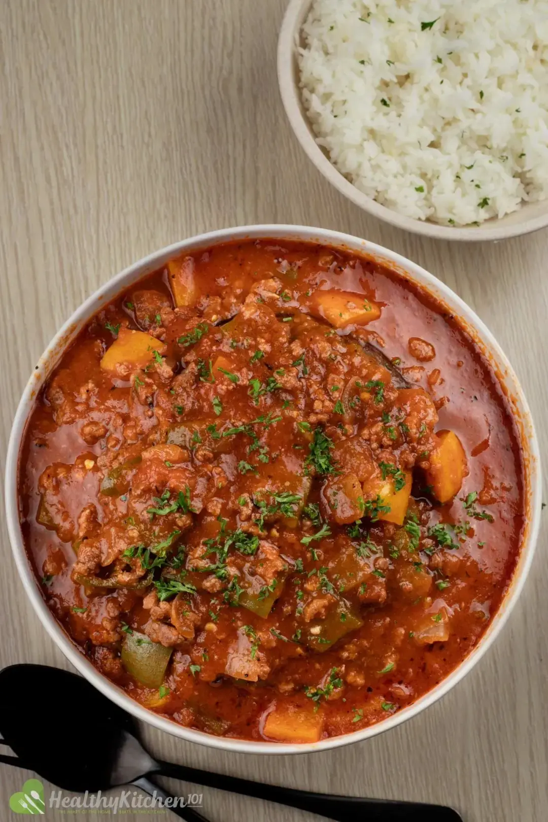 A soup bowl of sweet pepper, beef, and parsley, put next to dark utensils and cooked rice