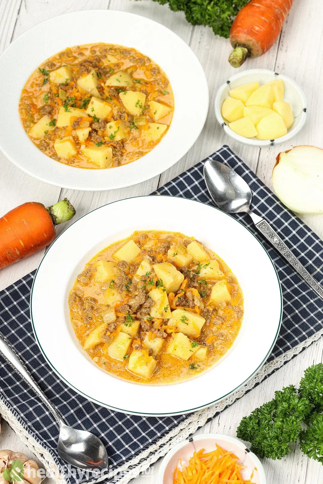 Two plates of cheeseburger soup laid on a blue tablecloth and near a spoon, fresh parsley, carrots, a small dish of julienned carrots, and a small dish of potato cubes