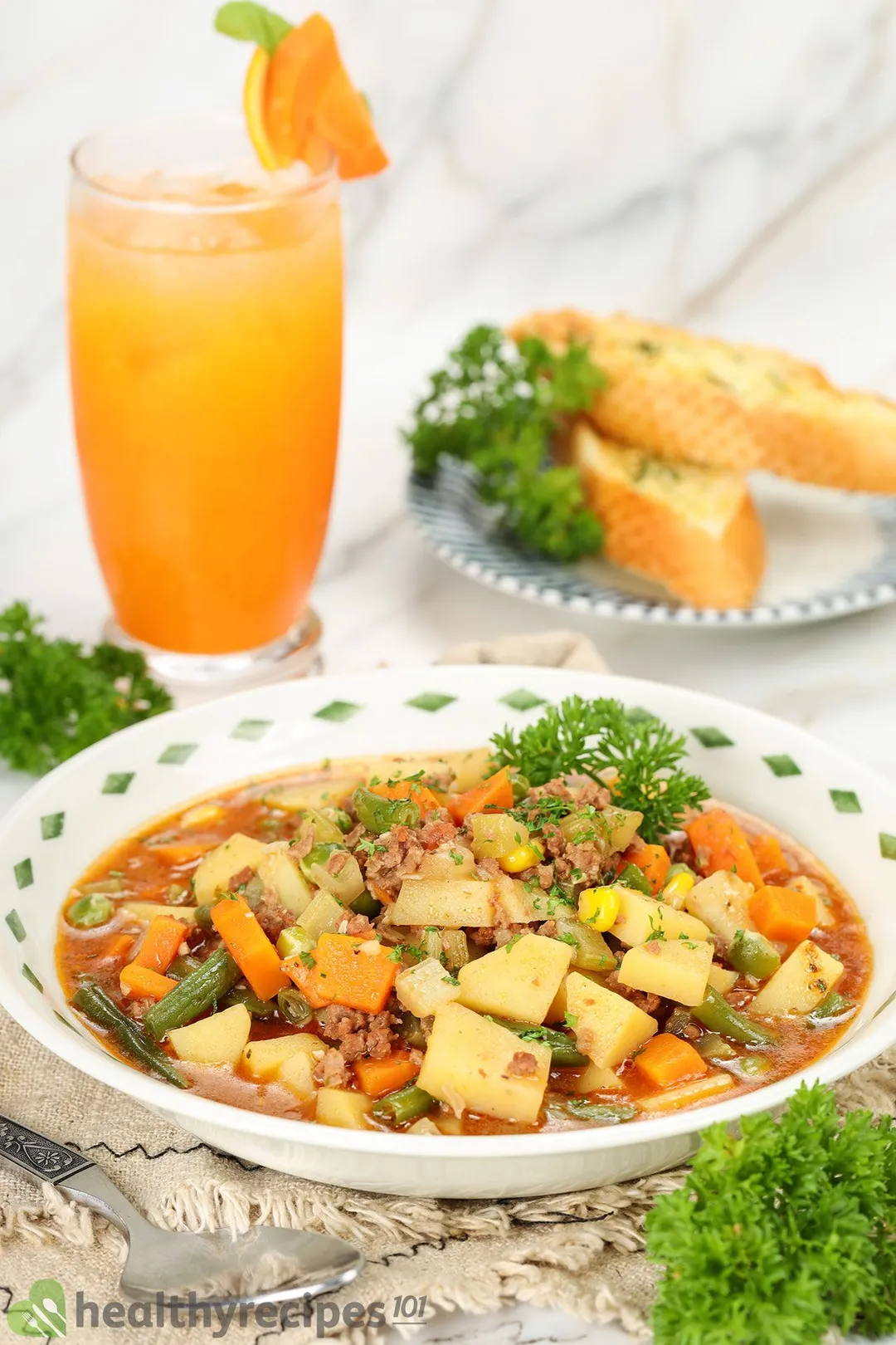 a plate of ground beef soup next to a glass of juice and a plate of garlic bread