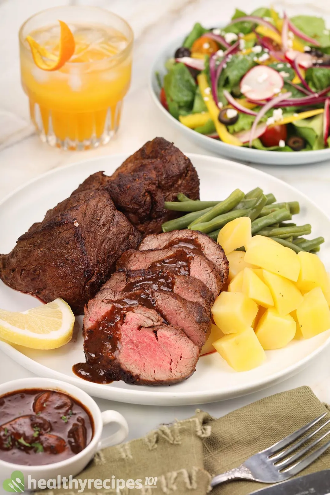 a plate of roasted beef tender loin with potato cubed, green beans decorated with a plate of salad and a glass of orange juice