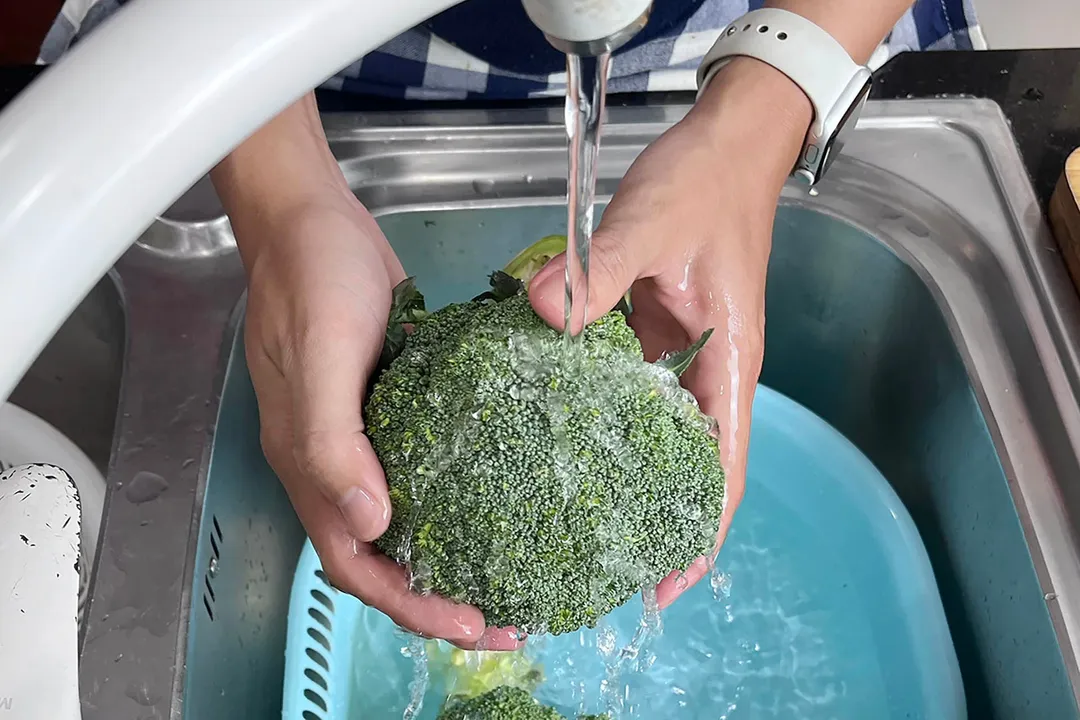 two hand washing a broccoli under a water dispenser