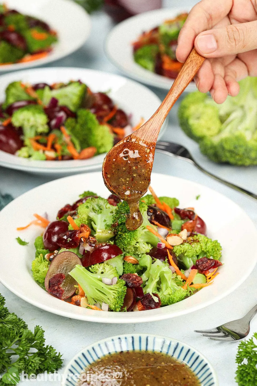 A hand using a wooden spoon to drizzle a dressing over plates of vegan broccoli salad.